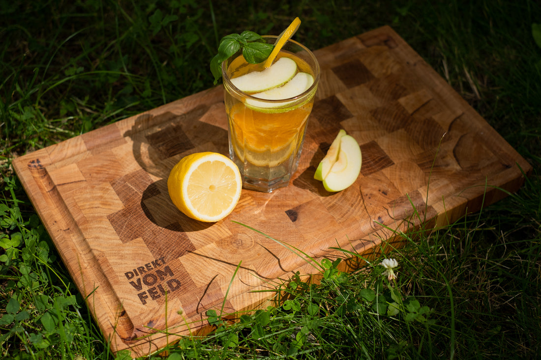 Sommerliche Kräutersirup-Limonade in einem hohen Glas mit mit Strohalm garniert mit Apfelscheiben und Zitronenhälfte auf einem Holzbrett im hohen Gras