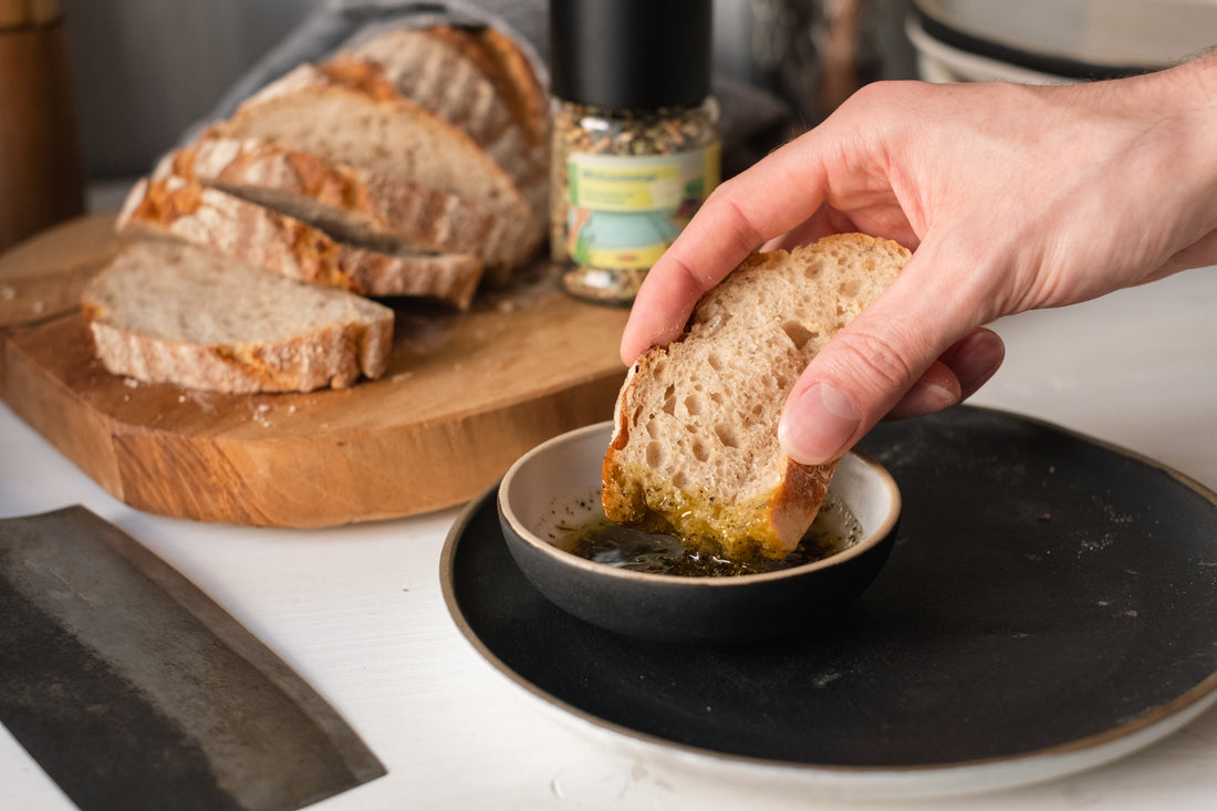 Eine Hand dippt eine Scheibe Brot in die Midsommar Vinaigrette in einer kleinen Schale, die auf einem dunklen Teller steht. Im Hintergrund ist das Brot zusehen mit der Bio Gewürzmischung Midsommar von Direkt vom Feld